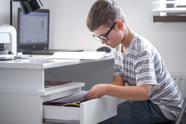 Kleiner Junge mit Brille sitzt an seinem Schreibtisch vor einem Laptop und sucht nach etwas in einer Schublade.