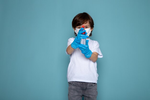 Kleiner Junge niedlich in den blauen Handschuhen und in der weißen sterilen Schutzmaske unter Verwendung des Sprays auf der blauen Wand