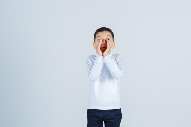 Kleiner Junge schreit oder kündigt etwas im weißen Hemd, in der Hose an und sieht aufgeregt aus. Vorderansicht.