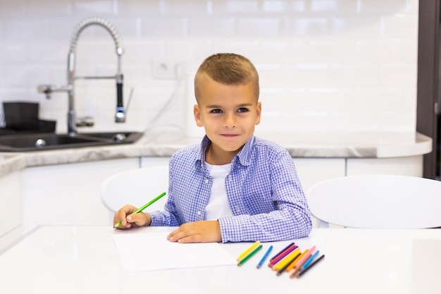 Kleiner Junge sitzt an einem Tisch in einer hellen Küche und zeichnet mit Bleistiften