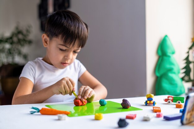 Kleiner kaukasischer Junge, der mit farbigem Plastilin spielt und Figuren auf dem weißen Tisch macht. Glückliche Kinderidee