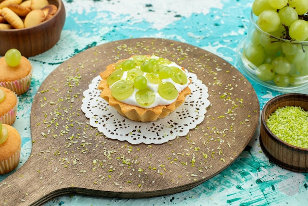 Kostenloses Foto kleiner leckerer kuchen mit köstlicher sahne und geschnittenen und frischen traubenplätzchen auf blaulichtschreibtisch