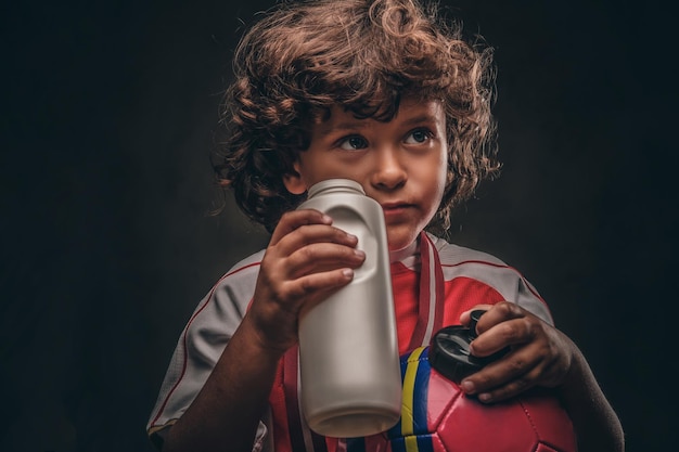 Kleiner Meisterjunge in Sportbekleidung hält einen Ball und trinkt Wasser aus einer Flasche. Getrennt auf dem dunklen strukturierten Hintergrund.