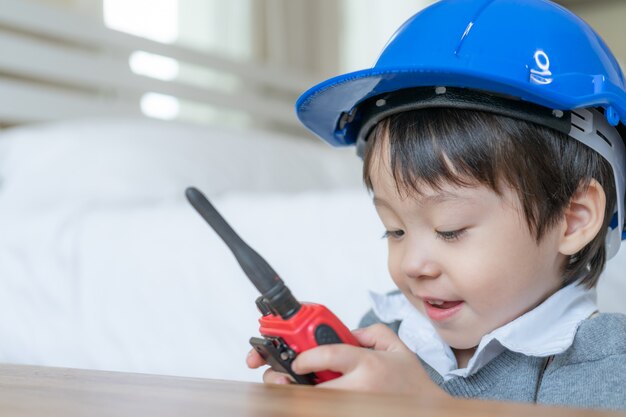 Kleiner netter Junge, der blauen Sturzhelm trägt und zur Unterhaltung mit rotem Funksprechgerät redio im Schlafzimmer genießt