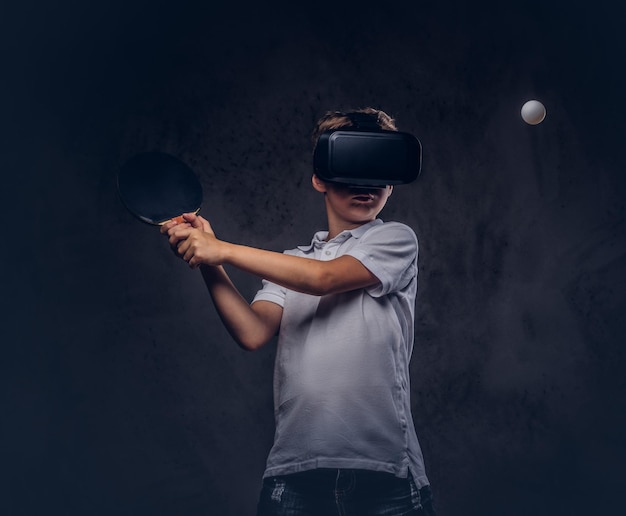 Kleiner rothaariger Junge in einem weißen T-Shirt, der mit einer Virtual-Reality-Brille Tischtennis spielt. Getrennt auf einem dunklen strukturierten Hintergrund.