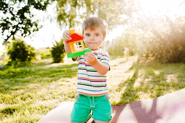 Kleiner süßer Junge, der ein Spielzeughaus im Garten hält und nachdenklich aussieht.