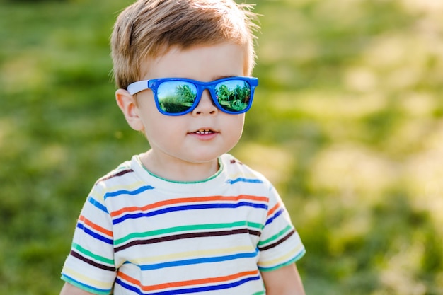 Kleiner süßer Junge, der im Garten in der hellen Sonnenbrille und im Lächeln bleibt.