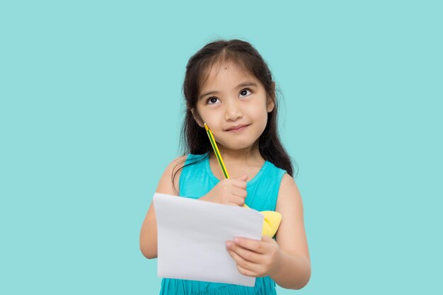 Kleines asiatisches Mädchen, das Papier und Bleistift in den Händen hat und den Schriftsteller nachschlägt, der auf Muse wartet, isoliert auf grünem Hintergrund