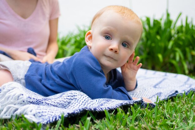 Kleines Baby, das auf Plaid liegt, Hand nahe Gesicht hält und wegschaut. Nahaufnahmeporträt im Garten. Junge Mutter sitzt. Sommer Familienzeit, sonnige Tage und Frischluftkonzept