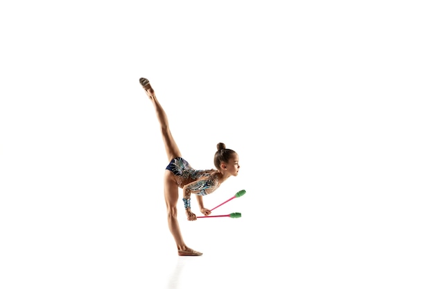 Kostenloses Foto kleines flexibles mädchen lokalisiert auf weißer wand. kleines weibliches modell als rhythmische gymnastikkünstlerin im hellen trikot. anmut in bewegung, action und sport. übungen mit den streitkolben machen.