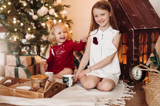 Kleines hübsches Mädchen mit gewelltem Haar sitzt mit einer Schachtel mit einem Geschenk und freut sich mit ihrer älteren Schwester