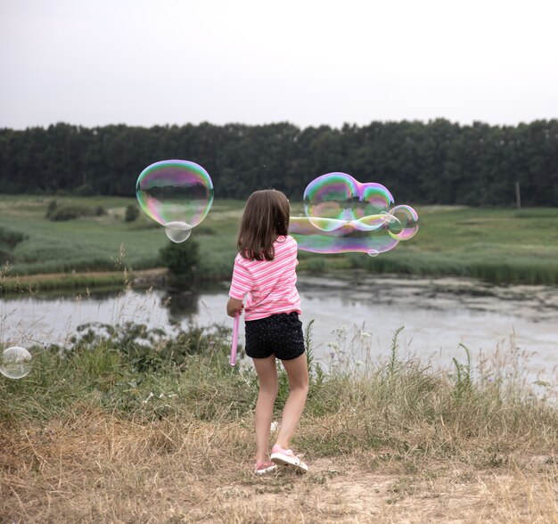 Kleines Kind Mädchen startet riesige Seifenblasen im Hintergrund schöne Natur, Rückansicht.