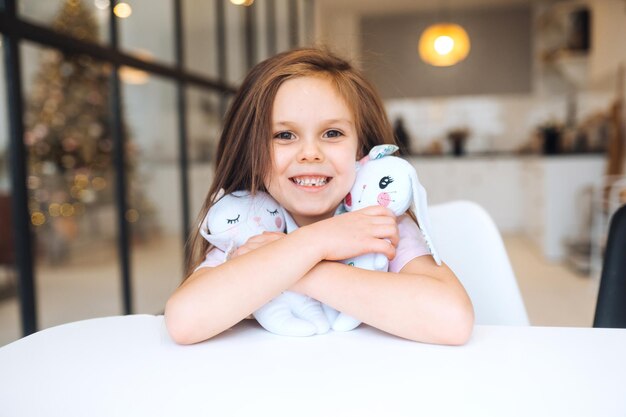 Kleines lächelndes Mädchen, das Lieblingsspielzeug umarmt, während es am Tisch sitzt
