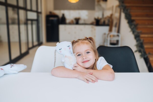 Kleines lächelndes Mädchen, das Lieblingsspielzeug umarmt, während es am Tisch sitzt