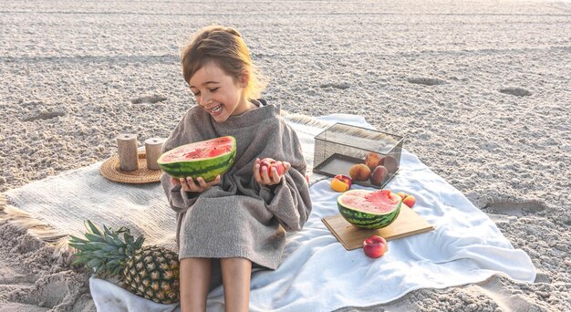 Kleines Mädchen an der sandigen Küste bei einem Picknick mit Früchten