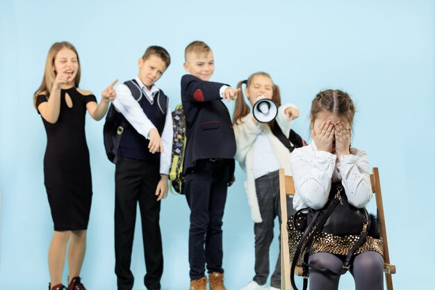 Kleines Mädchen, das allein auf Stuhl sitzt und einen Akt des Mobbings erleidet, während Kinder verspotten