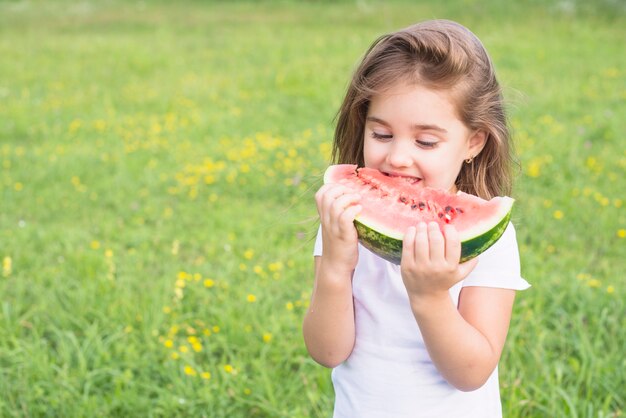 Kleines Mädchen, das auf dem Gebiet isst rote Scheibe der Wassermelone steht