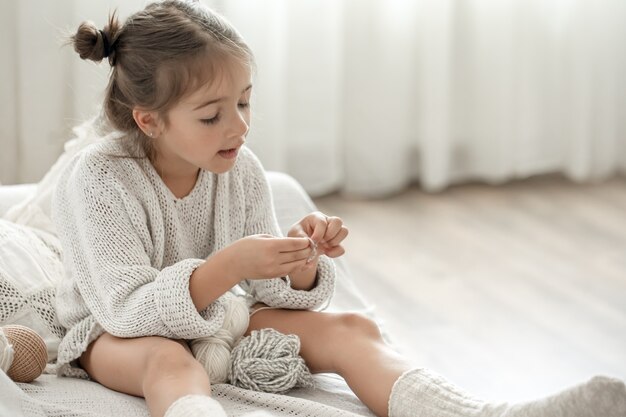 Kleines Mädchen, das auf dem Sofa sitzt und lernt, zu stricken, Freizeitkonzept zu Hause.