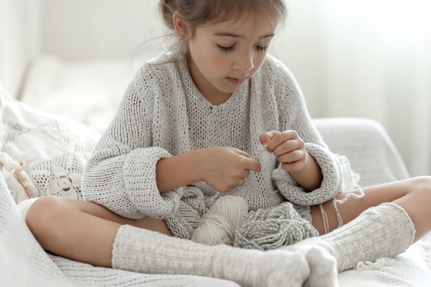 Kleines Mädchen, das auf dem Sofa sitzt und lernt, zu stricken, Freizeitkonzept zu Hause.