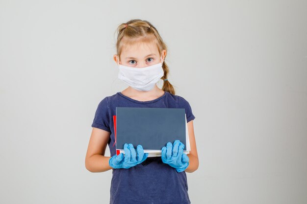 Kleines Mädchen, das Bücher und Notizbücher in der Vorderansicht des T-Shirts, der Handschuhe und der medizinischen Maske hält.