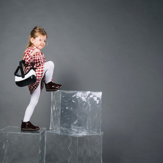 Kleines Mädchen, das die Gläser der virtuellen Realität klettern auf transparenten Blöcken gegen grauen Hintergrund hält