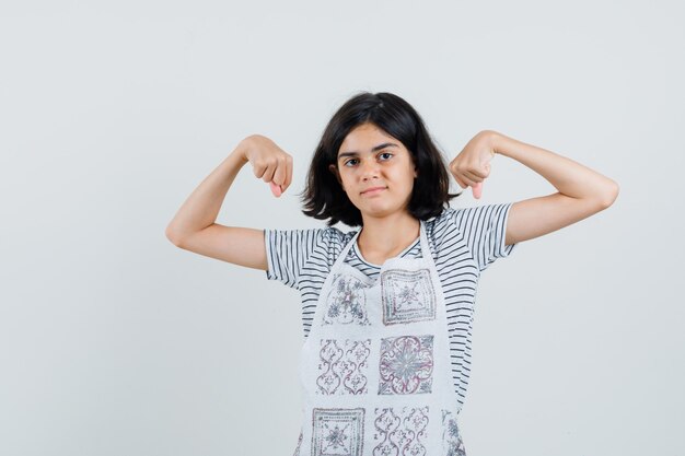 Kleines Mädchen, das die Muskeln der Arme im T-Shirt, in der Schürze zeigt und selbstbewusst aussieht