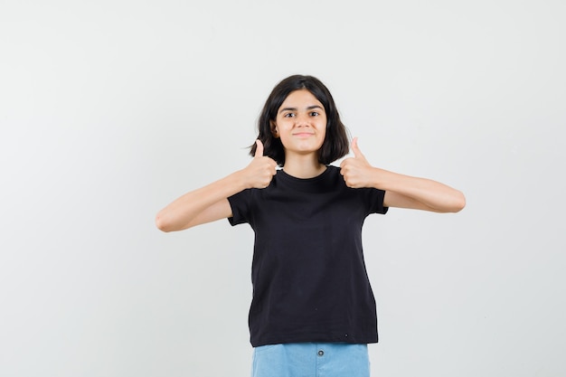 Kleines Mädchen, das doppelte Daumen in schwarzem T-Shirt, Shorts und selbstbewusstem Blick von vorne zeigt.