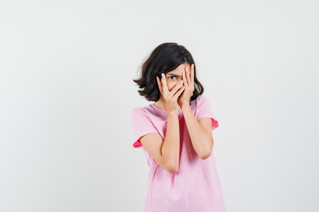 Kleines Mädchen, das durch Finger im rosa T-Shirt schaut und neugierig schaut, Vorderansicht.