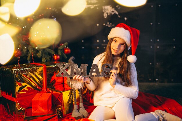 Kleines Mädchen, das durch Weihnachtsbaum sitzt und Geschenke auspackt