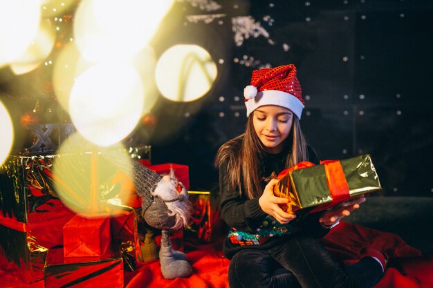 Kleines Mädchen, das durch Weihnachtsbaum sitzt und Geschenke auspackt