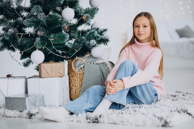 Kleines Mädchen, das durch Weihnachtsbaum sitzt