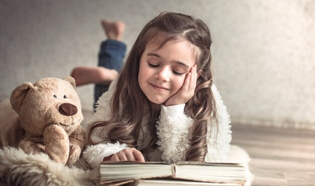 kleines Mädchen, das ein Buch mit einem Teddybär auf dem Boden liest, Konzept der Entspannung und der Freundschaft