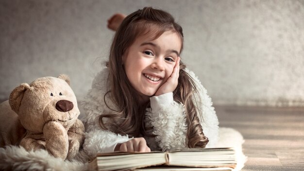 kleines Mädchen, das ein Buch mit einem Teddybär auf dem Boden liest, Konzept der Entspannung und der Freundschaft