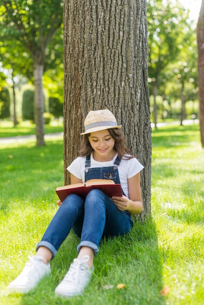 Kleines Mädchen, das ein Buch nahe bei einem Baum liest