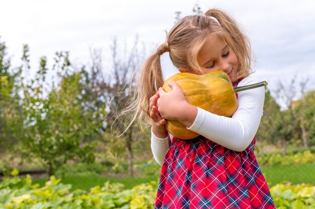 Kleines Mädchen, das ein Flanellkleid trägt und einen Kürbis auf einem Bauernhoffeld umarmt