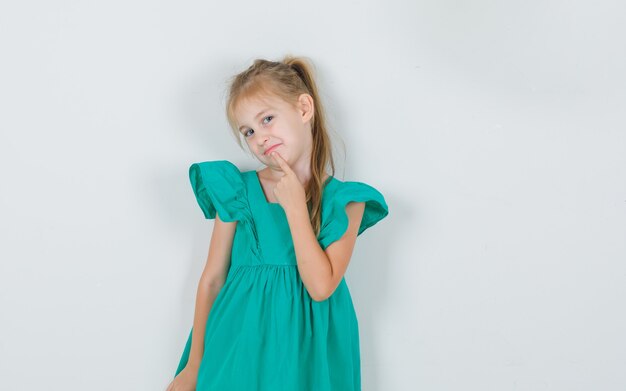 Kleines Mädchen, das Finger auf Kinn im grünen Kleid hält und hübsch aussieht. Vorderansicht.