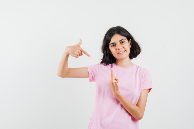 Kleines Mädchen, das Finger oben und unten im rosa T-Shirt zeigt und optimistisch, Vorderansicht schaut.