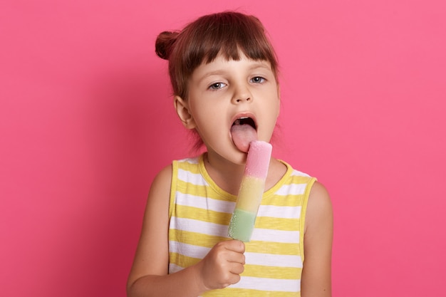 Kleines Mädchen, das Fruchteis isst, weit geöffneter Mund, gestreiftes Kleid tragend, hat dunkles Haar und zwei Knoten, posierend isoliert über rosa Wand.