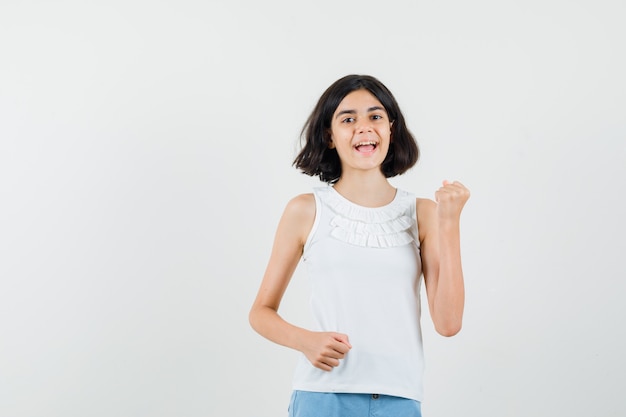 Kleines Mädchen, das geballte Faust in weißer Bluse, Shorts erhebt und glücklich schaut. Vorderansicht.