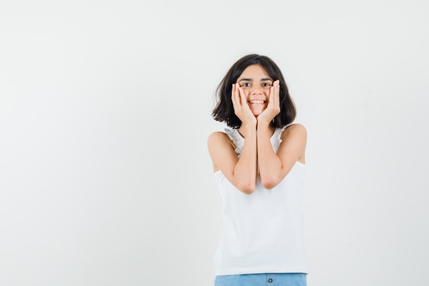 Kleines Mädchen, das Gesicht auf ihren Händen in der weißen Bluse, in den Shorts und im reizenden Aussehen kissen. Vorderansicht.