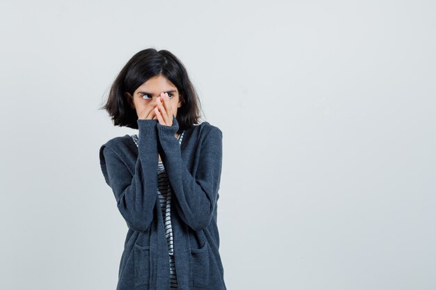 Kleines Mädchen, das Hände auf Gesicht in T-Shirt, Jacke hält und ängstlich schaut,