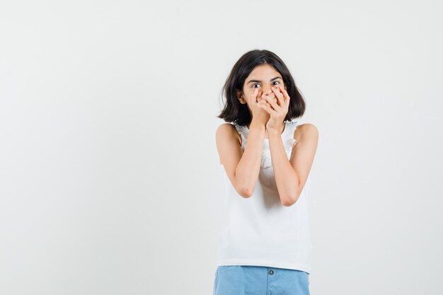 Kleines Mädchen, das Hände auf Mund in weißer Bluse, Shorts hält und ängstlich schaut, Vorderansicht.