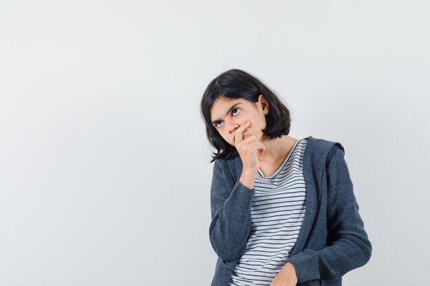 Kleines Mädchen, das Hand am Kinn im T-Shirt, in der Jacke hält und zögernd schaut