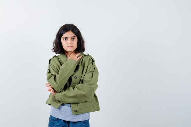 Kleines Mädchen, das Hand auf der Brust in Mantel, T-Shirt, Jeans hält und selbstbewusst aussieht. Vorderansicht.