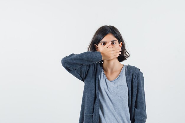 Kleines Mädchen, das Hand auf Mund in T-Shirt, Jacke hält und ruhig schaut.