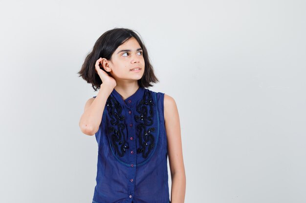 Kleines Mädchen, das Hand hinter Ohr in blauer Bluse hält und neugierige Vorderansicht schaut.