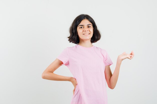 Kleines Mädchen, das Handfläche im rosa T-Shirt beiseite spreizt und fröhlich aussieht. Vorderansicht.