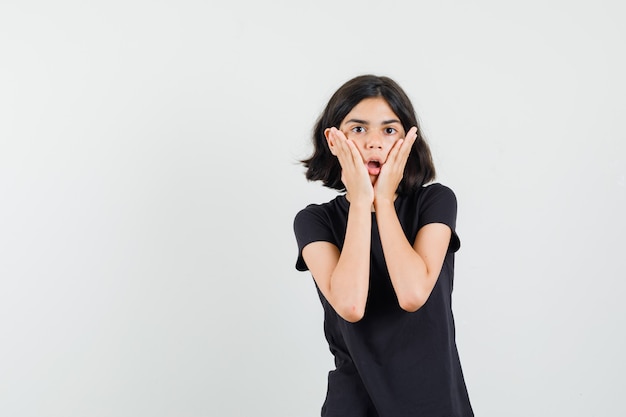 Kleines Mädchen, das Handflächen auf Wangen im schwarzen T-Shirt hält und überrascht schaut. Vorderansicht.