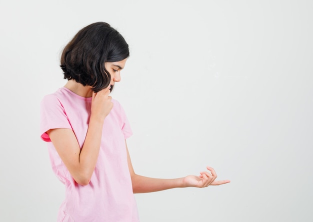 Kleines Mädchen, das ihre Handfläche im rosa T-Shirt betrachtet und nachdenklich schaut. Vorderansicht.