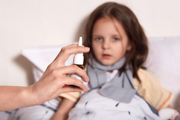 Kleines Mädchen, das im Bett liegt, ihre Mutter, die ihre laufende Nase mit Nasenspray behandelt, dunkelhaariges weibliches Kind, das Kamera betrachtet